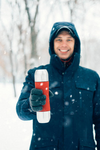 快乐的人拿着热水瓶在冬天的公园里下雪。 冬天在森林里喝热饮料的年轻人