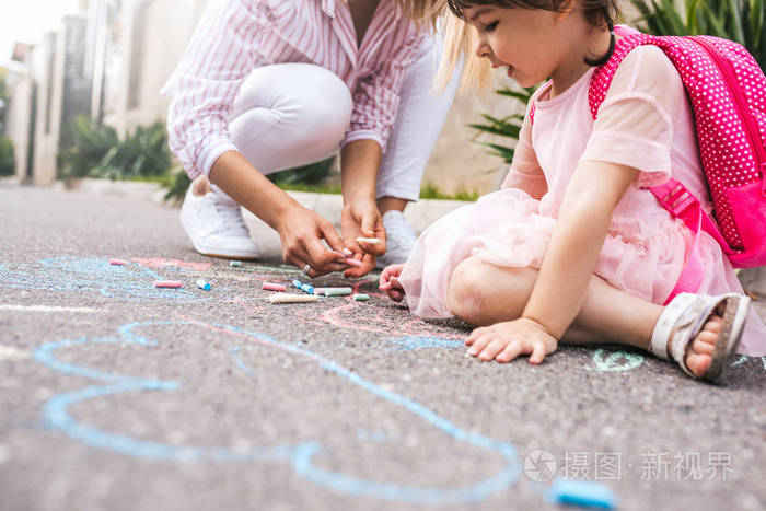 可爱的小女孩和妈妈在人行道上画着五颜六色的粉笔画。 高加索金发女性和她的孩子学龄前儿童一起玩背包户外。 母亲和儿童活动