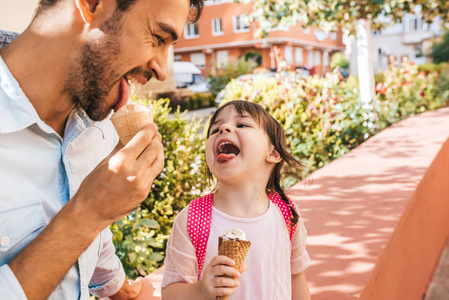 特写水平肖像可爱的小女孩坐在城市街道上和爸爸一起在户外吃冰淇淋。 快乐的女孩穿着粉红色的裙子和父亲在一起。 快乐的学龄前儿童