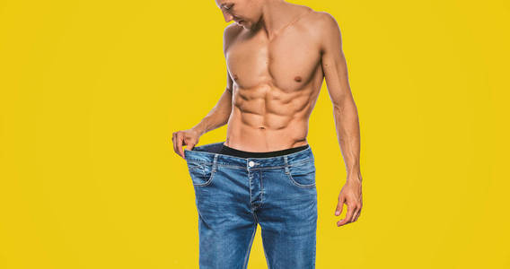 水平工作室形象的年轻白种人适合男子穿着大蓝色牛仔裤后，饮食显示的躯干。 减肥后有吸引力的男人在黄色背景上摆姿势。 复制空间。