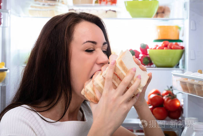 一位饥饿的年轻女子在冰箱旁吃三明治