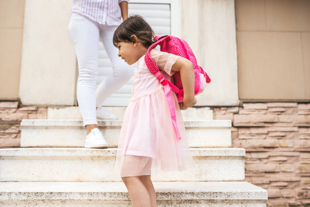 母亲站在楼梯上，靠在家里的户外，在女儿离开幼儿园时向她道别。 可爱快乐的小女孩穿着背包去幼儿园。 教育