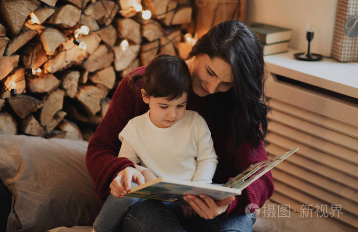 漂亮的母亲和她的小女儿晚上在家看书,装饰着舒适和圣诞节.