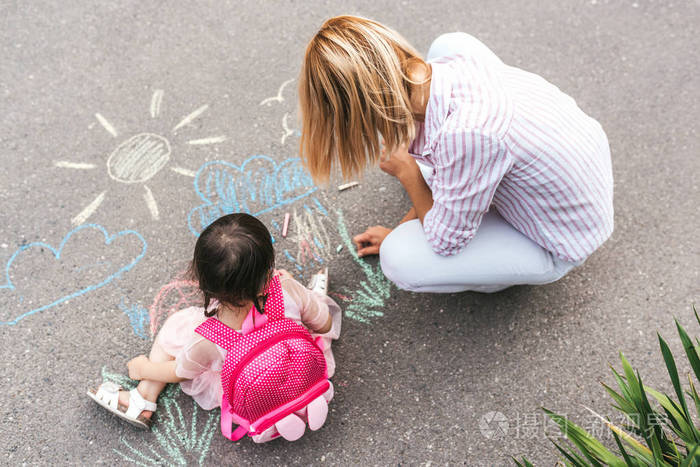 可爱的小女孩和她的母亲在人行道上画着五颜六色的粉笔。 高加索金发女性和她的孩子学龄前儿童一起玩背包户外。 母亲和儿童活动