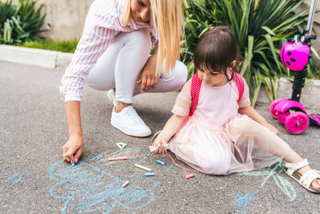 幸福的小女孩和母亲在人行道上用粉笔画画的水平形象。 白种人女性与儿童学龄前儿童一起玩背包户外。 妈妈和孩子的活动。 良好的关系