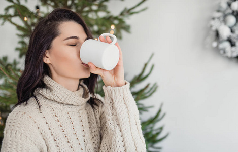 一幅美丽的年轻女子在家中摆姿势，在圣诞树旁边喝着一杯热饮料的肖像。白人女性在客厅里放松，在圣诞节装饰上喝杯咖啡