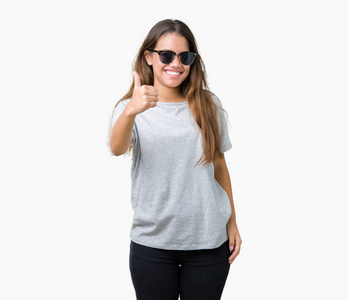 年轻漂亮的黑发女人戴着太阳镜，在孤立的背景，用手做快乐的竖起大拇指的手势。 赞许的表情看着相机，显示出成功。