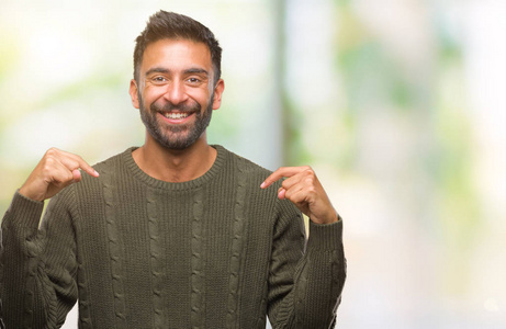 成年西班牙裔男子穿着冬天的毛衣，在孤立的背景下，自信地微笑着，用手指指着自己，骄傲和快乐。