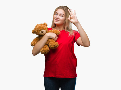 年轻的白种人女人抱着可爱的泰迪熊在孤立的背景下做OK标志与手指优秀的符号