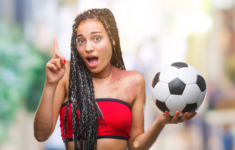 年轻的留着辫子的非裔美国人，有出生标记，在孤立的背景下拿着足球，惊讶地发现了一个想法或问题，指着一个快乐的脸第一