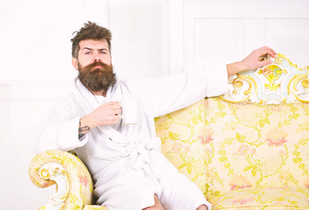 胡子和胡须的男人坐在豪华沙发上享受早晨。精英休闲理念。男子在昏昏欲睡的脸上浴袍, 喝咖啡, 在豪华酒店在早上, 白色背景