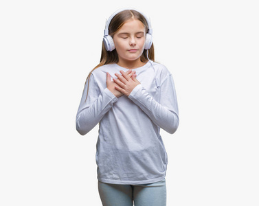 年轻漂亮的女孩戴着耳机听音乐，在孤立的背景上微笑，双手放在胸部，闭着眼睛，脸上有感激的手势。 健康概念。