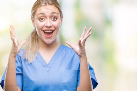 年轻的金发外科医生女医生穿着医疗制服，在孤立的背景下庆祝疯狂，并惊讶于成功，举起手臂和睁开眼睛尖叫兴奋。 赢家概念