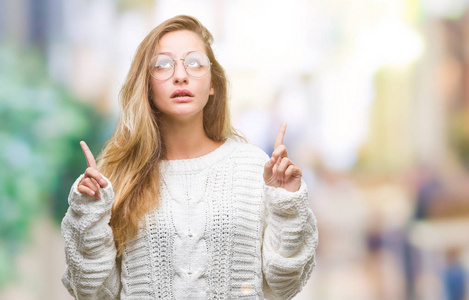 年轻漂亮的金发女人穿着冬天的毛衣和太阳镜，在孤立的背景上惊讶和惊讶地抬头，用手指和举起的手臂指着。