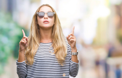 年轻漂亮的金发女人戴着太阳镜在孤立的背景上惊讶和惊讶地抬头，用手指和举起的手臂指着。