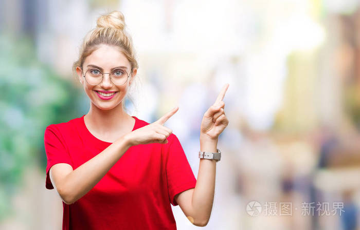年轻漂亮的金发女人穿着红色的t恤和眼镜，在孤立的背景上微笑，看着相机，双手和手指指向一边。