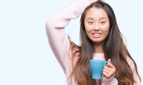 年轻的亚洲女人喝咖啡在孤立的背景下，压力与手在头上，震惊与羞愧和惊讶的脸，愤怒和沮丧。因为错误而恐惧和不安。