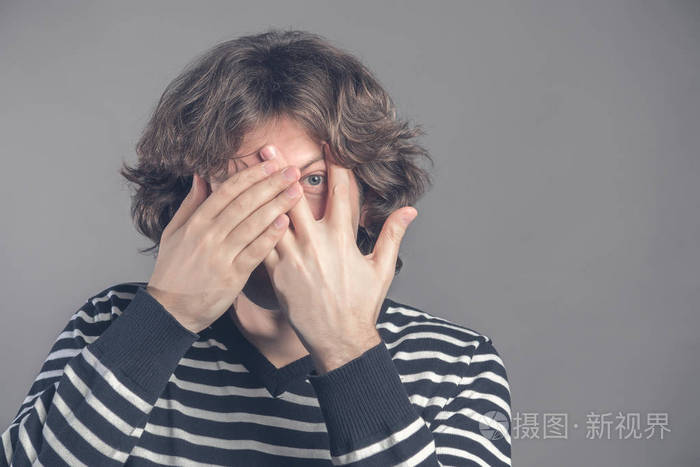 英俊的男人深色卷发刚毛隐藏着脸双手透过手指窥视穿着条纹毛衣在灰色