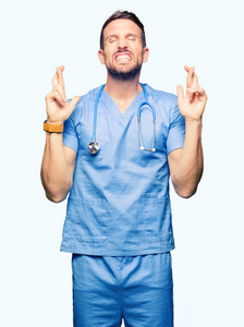 英俊的医生穿着医疗制服，在孤立的背景上微笑着交叉手指，带着希望和眼睛闭上。 运气和迷信的概念。