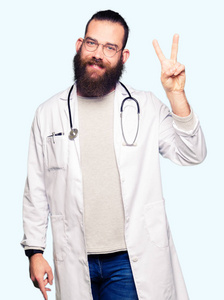 年轻的金发医生，留着胡子的男人，穿着医疗外套，用手指举着第二根手指，微笑着自信和快乐。