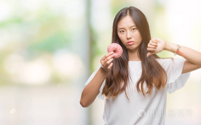 年轻的亚洲女人吃甜甜圈在孤立的背景和愤怒的脸消极的迹象显示不喜欢拇指向下拒绝的概念