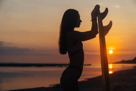 剪影苗条的女孩与冲浪板在手在海滩在美丽的日落背景