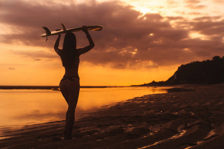 剪影苗条的女孩与冲浪板在手在海滩在美丽的日落背景