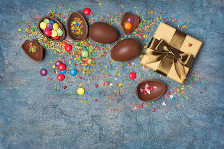 顶部观看巧克力传统复活节彩蛋和金色礼品盒与明亮的彩色悬垂糖洒在蓝色混凝土背景与复制空间。