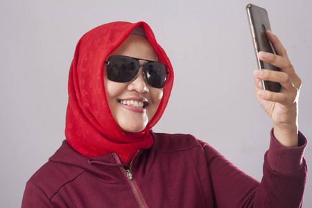 穆斯林女士戴着黑色太阳镜，穿着红色西装，戴着头巾，微笑着，在智能手机上拍摄照片