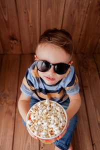 小可爱的孩子，小男孩，2，3岁的3D电影院眼镜，拿着水桶吃爆米花，在木制背景下吃快餐。 儿童童年生活方式的概念。 复制空间。