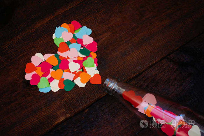 情人节创意DIY工艺礼品卡的想法。 许多五颜六色的纸心是从玻璃透明瓶在木制背景上用红色丝带。 爱情浪漫手工制作