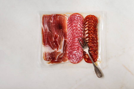 在打开的塑料包装中，用叉子在白色大理石背景上打开Jamon Salami Chorizo香肠切片的反胃肉盘。 平躺空间