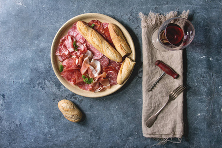在陶瓷盘子里，用面包和一杯红酒在蓝色纹理背景上的布上，用切好的Jamon Salami Chorizo香肠切片的反胃肉盘。 平躺