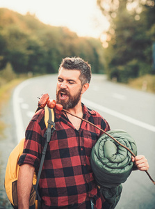 男子徒步者在棍子上吃香肠。带着食物在路上徒步的胡子人。带着背包和睡袋的希普斯特。游客在暑假旅行和徒步旅行。饮食理念
