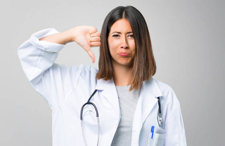 医生妇女听诊器显示拇指向下标志，表情阴性。 灰色背景的悲伤表情