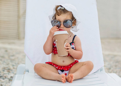快乐的女婴坐在带太阳镜的躺椅上，拿着饮料