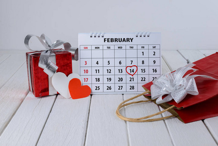 日历页红色手写心突出2月14日圣瓦伦日与红色礼品袋盒和心形在白色木桌上
