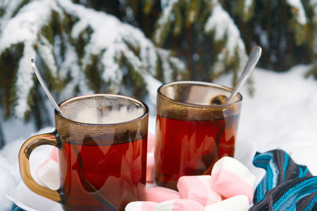 在冬天的雪地里，一棵树下裹着蓝色围巾，放着棉花糖的两杯茶