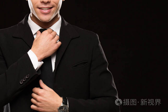 一个英俊的男子穿着优雅的黑色西装,微笑着调整他在黑色背景上的领带
