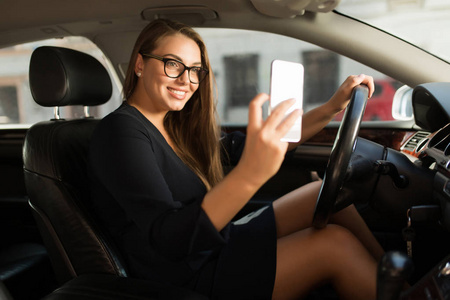 年轻漂亮的微笑女人，穿着黑色连衣裙和眼镜，坐在车里的方向盘后，愉快地用手机拍照