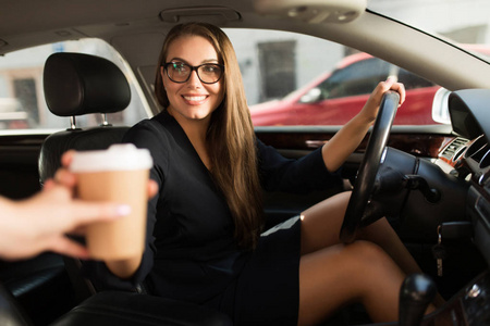 年轻漂亮的微笑着的女人，穿着黑色连衣裙，戴着眼镜，坐在车轮后面，坐在车里，高高兴兴地拿着咖啡去了