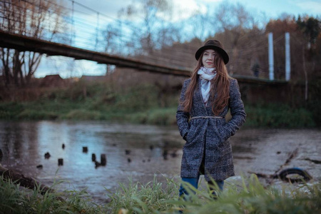 一个女孩站在河边，在一个阴天的晚上靠近桥。穿外套戴帽子的年轻女子站在水边
