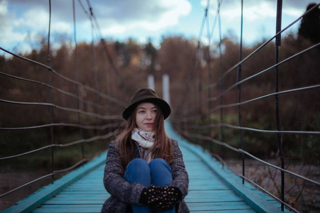村里一座桥上的年轻女子。 在秋天的阴天，穿着外套和帽子的女孩在木桥上散步