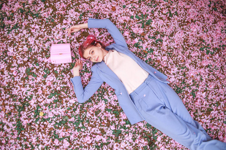 美丽的少女，穿着蓝色西装，粉红色的头发躺在草地上，花园里满是粉红色的花瓣