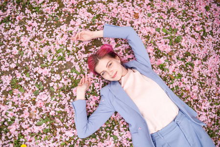 美丽的少女，穿着蓝色西装，粉红色的头发躺在草地上，花园里满是粉红色的花瓣