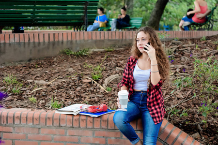 秋天，带着电话和一次性咖啡杯坐在公园里的年轻学生女孩。