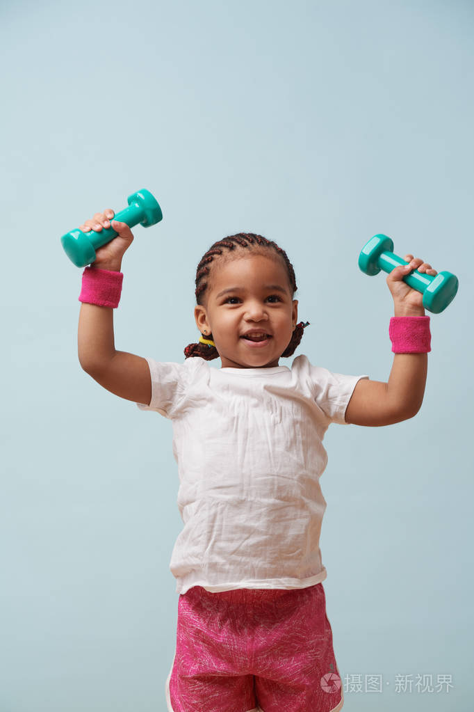 可爱的小女孩在淡蓝色背景下举起小彩色哑铃的肖像.
