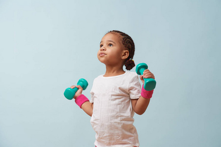可爱的小女孩在淡蓝色背景下举起小彩色哑铃的肖像。 粉红色短裤和腕带。 健身锻炼。