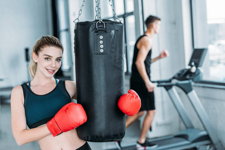 戴着拳击手套拿着拳击袋在健身房里对着镜头微笑的漂亮运动女孩