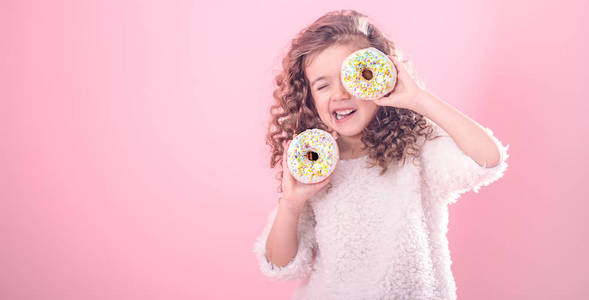 一个留着卷发手里拿着两个开胃甜甜圈的微笑小女孩的肖像，用粉红色背景上的甜甜圈闭上眼睛，一个发短信的地方